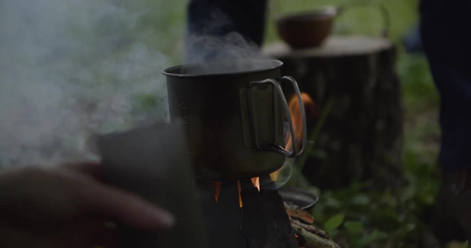 A dark metal camping mug with a wire handle sits over a small visible flame and emits a plume of steam with a person hands in the front and another one legs in the background on a grassy plain setting