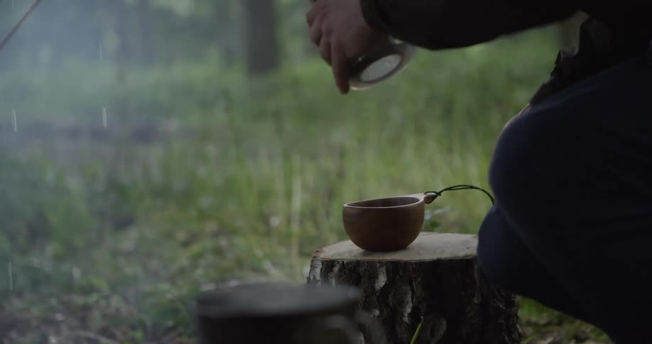 At a misty forest campsite a hand pours a hot drink into a wooden cup on a stump beside a kettle