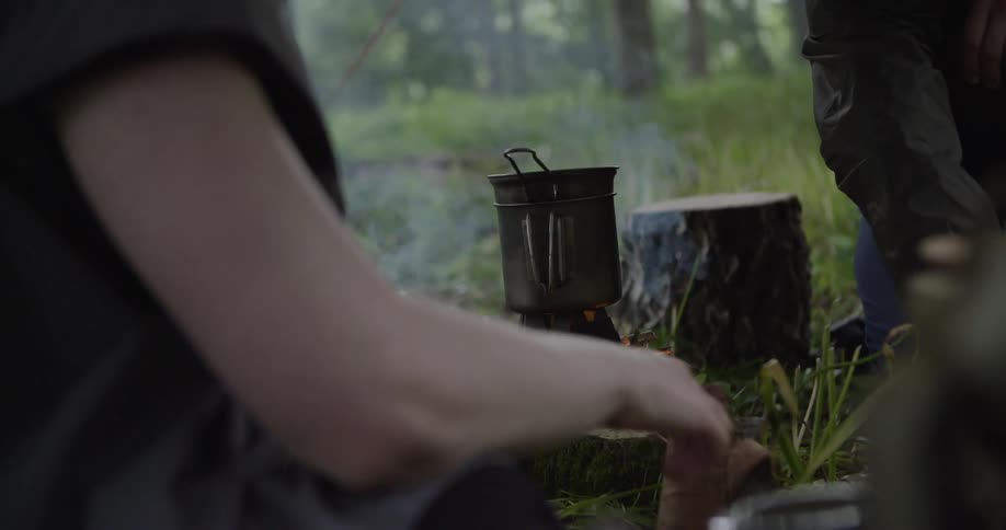 Two people sit beside a campfire in the woods at midday surrounded by tall trees as smoke drifts into the clear sky while they cook a meal over the fire