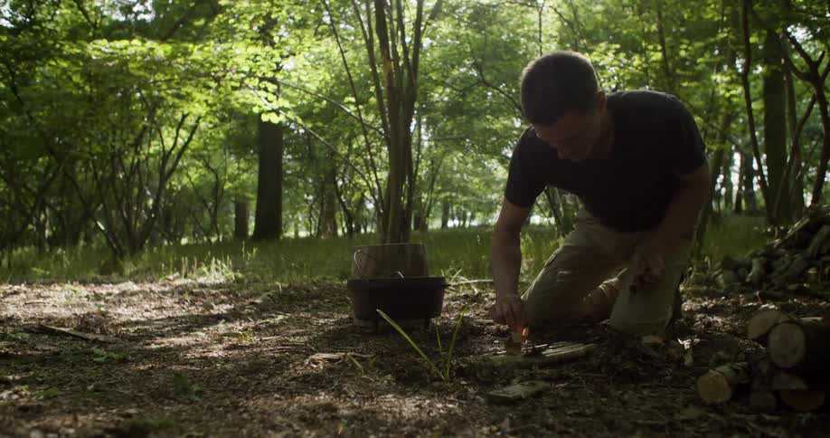 A young man wearing a dark shirt and tan pants kneels on the forest floor next to a pile of wood and a black pot attempting to start a small fire with tinder in a sunny clearing