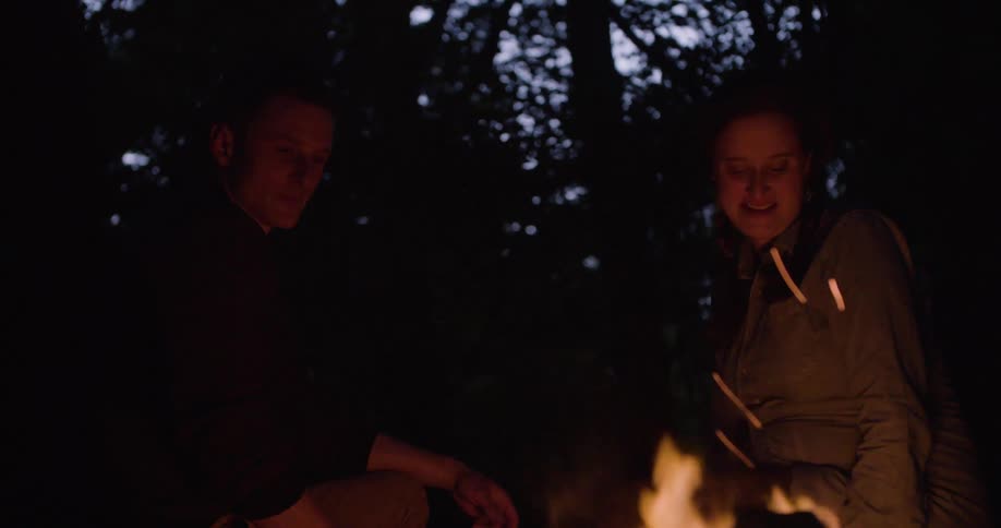 A young man and woman discussing while sitting close to a campfire in a woodland at night
