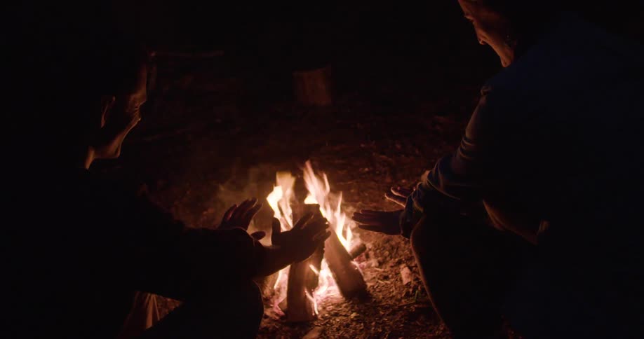 Two people warm their hands at the campfire at night