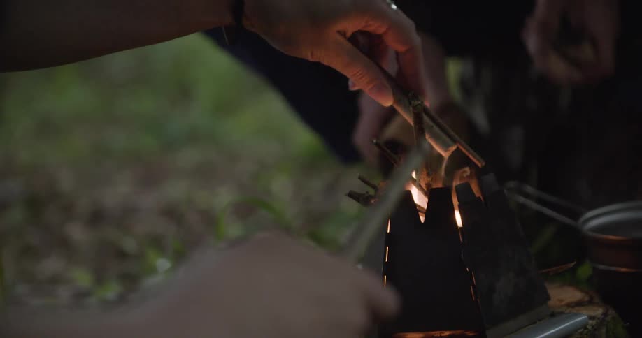 Two hands are actively placing small pieces of wood into the vertical opening of a pyramidshaped black metal portable stove that contains a burning fire in an outdoor setting