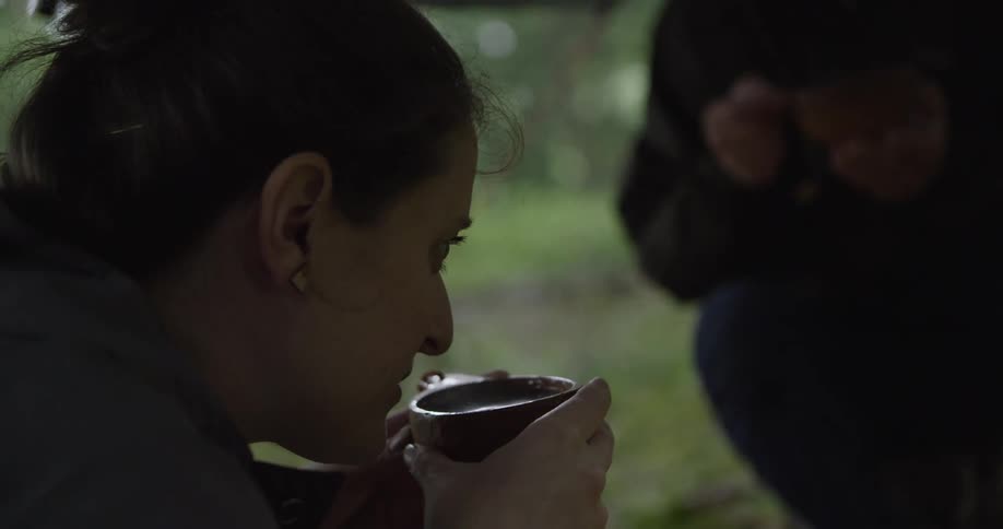 A woman is shown in a close side profile holding a rounded bowl with both hands and drinking from it while another person out of focus stands in the right background in a rainy outdoor environment
