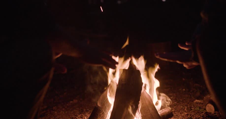 Two pairs of handsnear a brightly burning fire composed of large pieces of wood and glowing embers illuminating the immediate area against a completely dark background while sparks fly upward