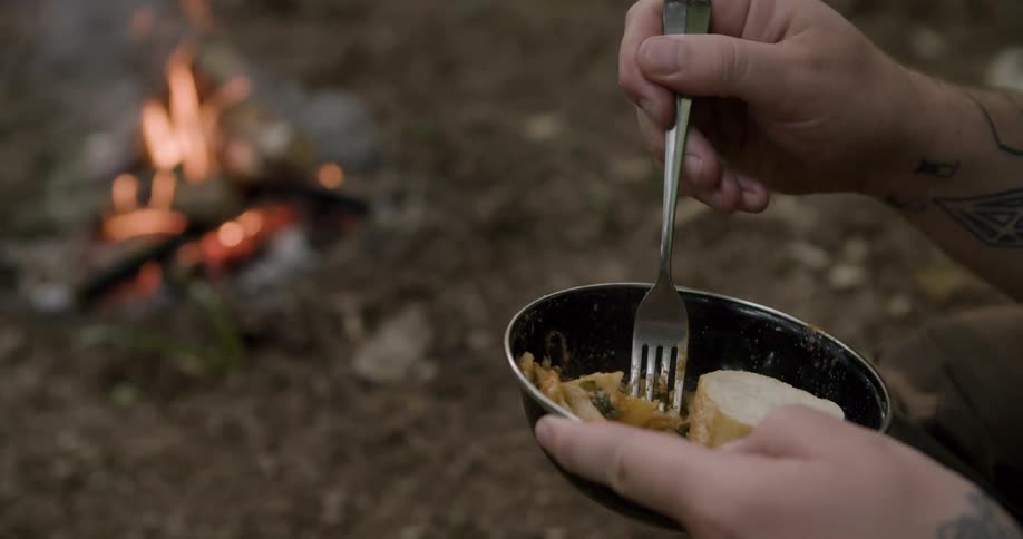 The man is mixing the food in his bowl by the campfire