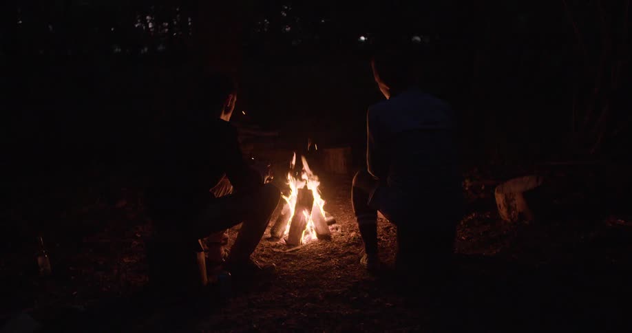 The silhouettes of two people are seen from the back sitting close to a bright crackling campfire in a dark forest setting at night with sparks flying from the flames