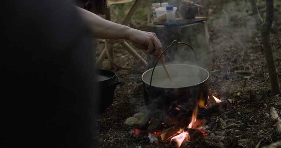 Person stirring the contents of a pot over an open fire in an outdoor setting