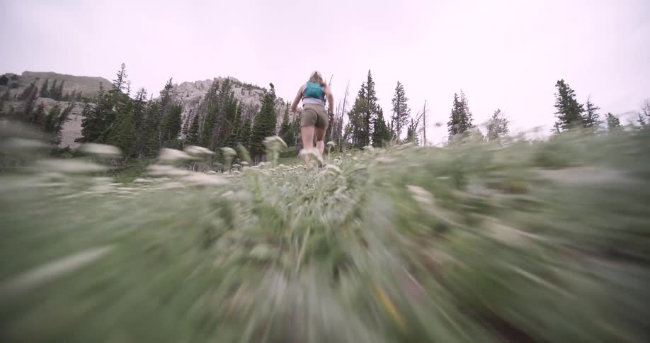 A dynamic lowangle view shows a woman wearing a tank top and shorts with a backpack running quickly uphill through long green grass toward a line of pine trees and rocky mountains under a cloudy sky