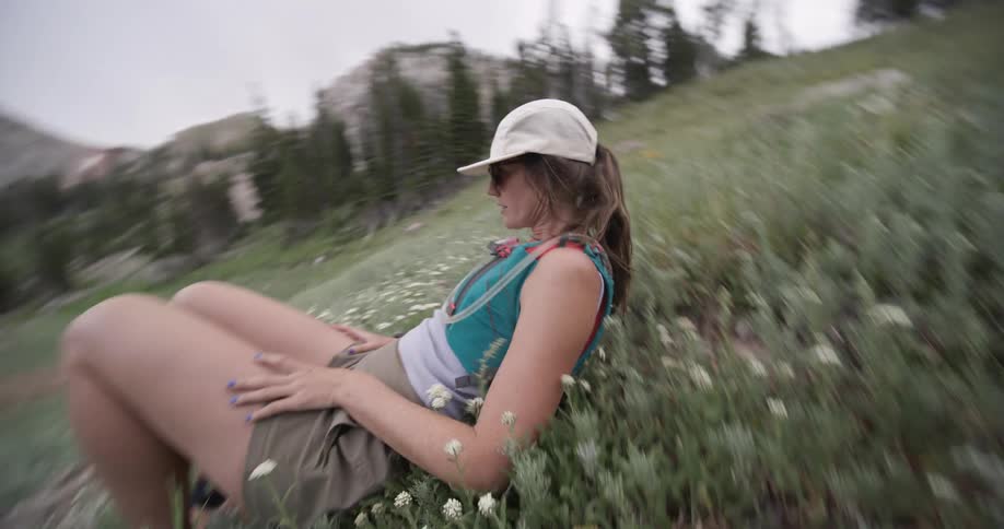 A person wearing a cap sunglasses and a hydration pack lies down in a grassy field with small white flowers looking up towards the sky with the surrounding mountain scenery out of focus