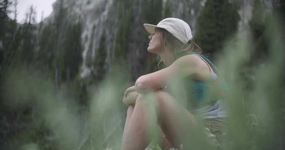A young woman sits on the forest floor at midday while hiking wearing a baseball hat as sunlight filters through the trees