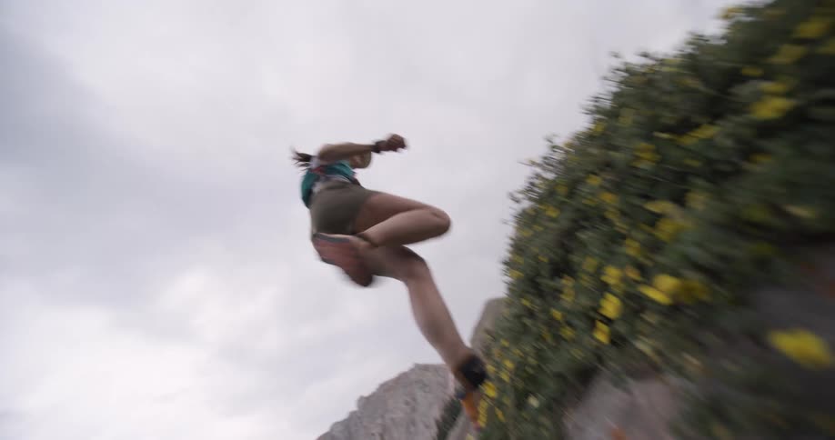 A person wearing running gear and a cap appears from the top right and performs a large running jump off a rocky ledge toward the lowerleft moving past a bank of grass and flowers framed by the rough terrain and an overcast sky