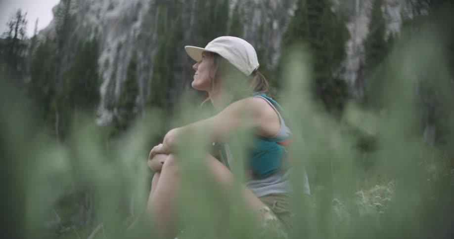 A woman sits with their legs drawn up and arms wrapped around them wearing a cap and tank top set against a blurry backdrop of a steep treecovered mountain slope