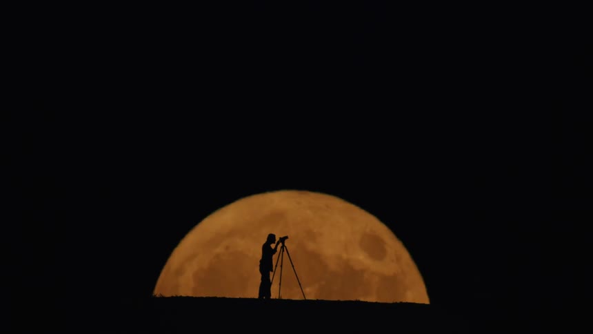 Person using a telescope is silhouetted by a large red moon at night