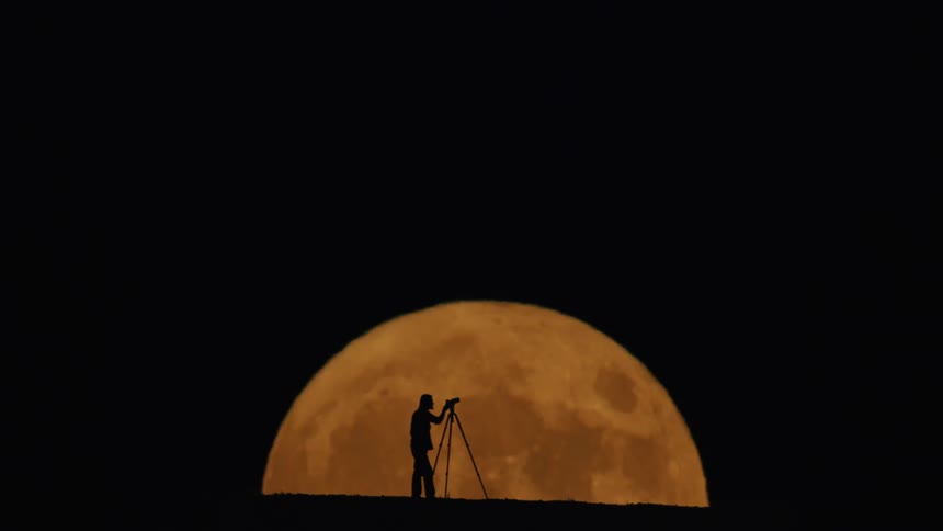 Silhouette of a photographer on a mountain with the reddish moon beside it at night