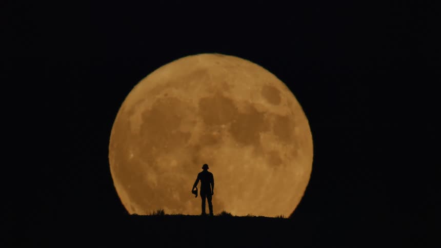 Silhouette of a man taking off his hat while standing on a hilltop facing a large full moon at night