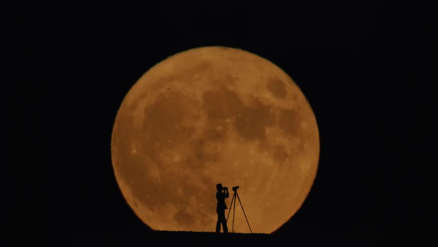 Person standing next to a tripod observing the orange full moon in the night sky