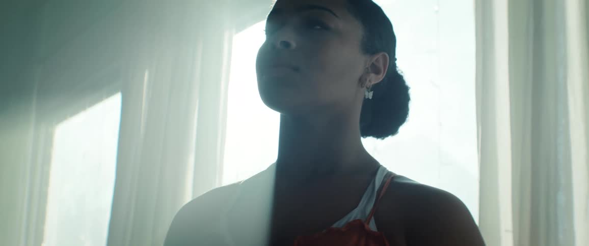A close profile view shows a young confident and determined woman with dark hair tied back wearing a light top looking upward with a determined expression in an intensely backlit room with a bright window and lens flare