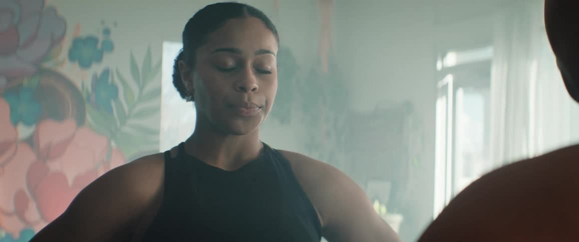 A close view shows a young woman with dark hair tied back wearing a black tank top closing her eyes to take a deep breath and then looking forward in a bright room with a painted floral wall