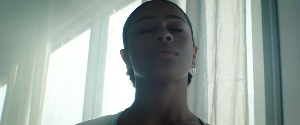 A close profile view shows a young woman with dark hair tied back and small butterfly earrings standing thoughtfully against a bright windowlit background creating a strong lens flare and silhouette effect