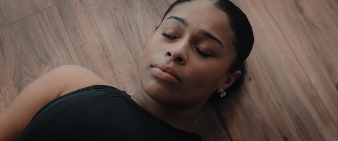 A close highangle view shows a young determined woman with dark hair pulled back wearing a black tank top resting with a tired but contemplative expression while lying on a wooden floor