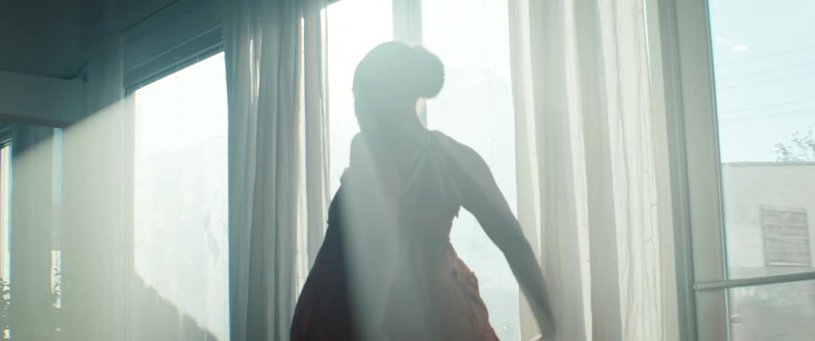 A silhouette of a young woman in a flowing orange dress is seen performing a ballet or dance move with a raised leg against large windows covered by sheer white curtains and intense backlighting