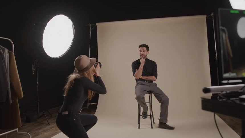 A young female photographer taking photos of a young man sitting on a stool in a photo studio