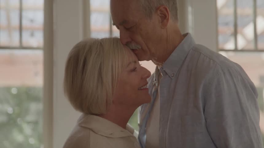 An elderly couple dances indoors at midday holding hands as the man kisses the woman on the forehead while she smiles