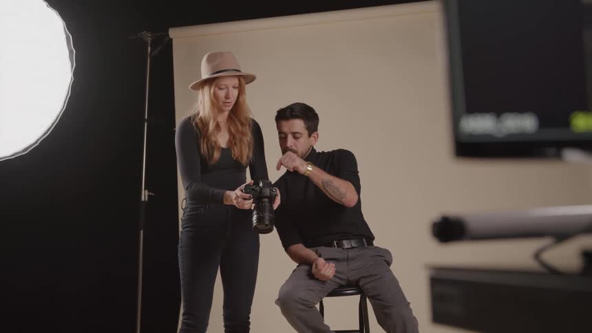 A young woman wearing a hat shows a man sitting on a stool the photograph she captured on a digital camera inside the studio