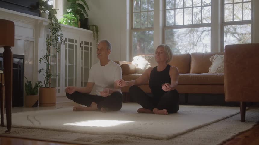 Man and woman sitting on yoga position over a carpet on a living room indoors during day time