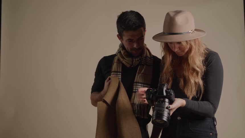 A man and a woman stand close together in an indoor space with a plain background with the man holding a brown coat and the woman holding a digital camera