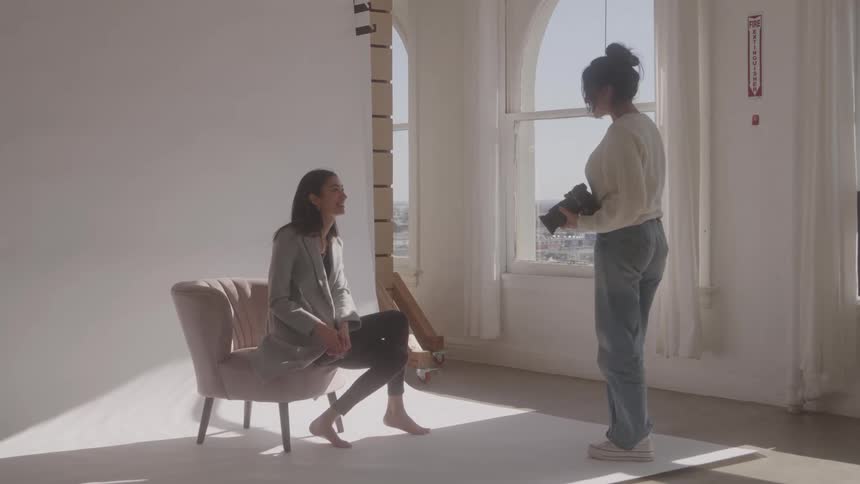 Female photographer standing up with a camera in her hand while talking to a female model sitting on a chair indoors during day time