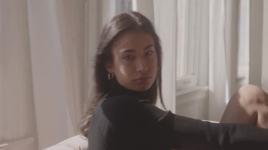A woman with long dark hair and wearing a black turtleneck sweater is seated indoors in front of a window covered by lightcolored curtains