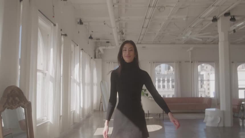 A young woman in a short black dress struts down a studio apartment towards a sofa passing a row of windows with white curtains then turns smiles and without ever stopping turns back in her original direction