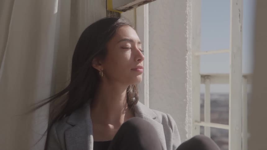 White curtains move with the wind as does the long hair of a young woman wearing a gray coat and a black top sitting on the window sil with her eyes closed
