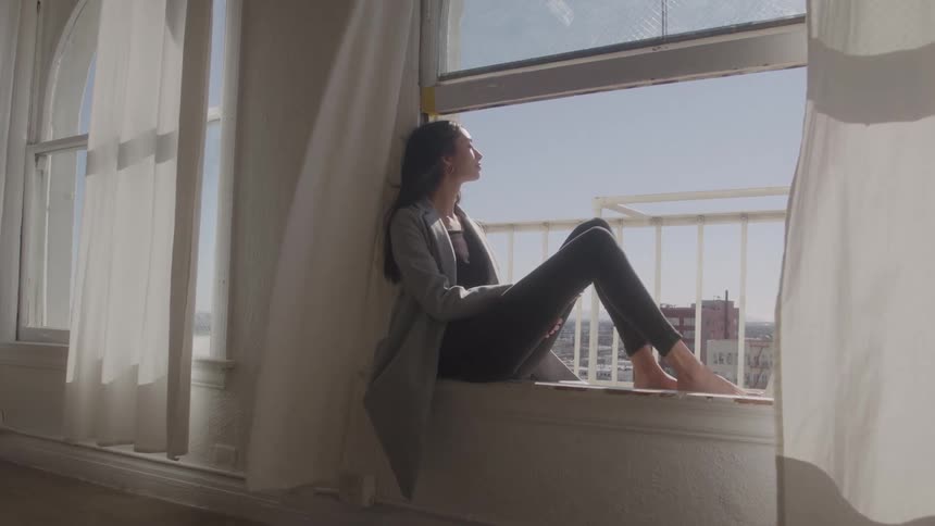 A young woman with long dark hair wears a light grey cardigan and dark pants while sitting barefoot on a wide sunlit windowsill looking contemplatively at the urban skyline outside