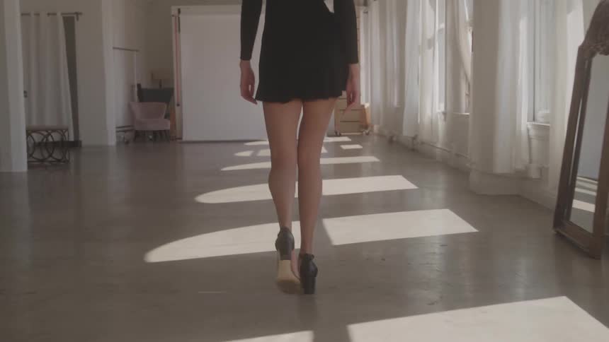 A woman with long bare legs and wearing a short black dress and highheeled shoes walks directly away from the camera across a large lightcolored floor