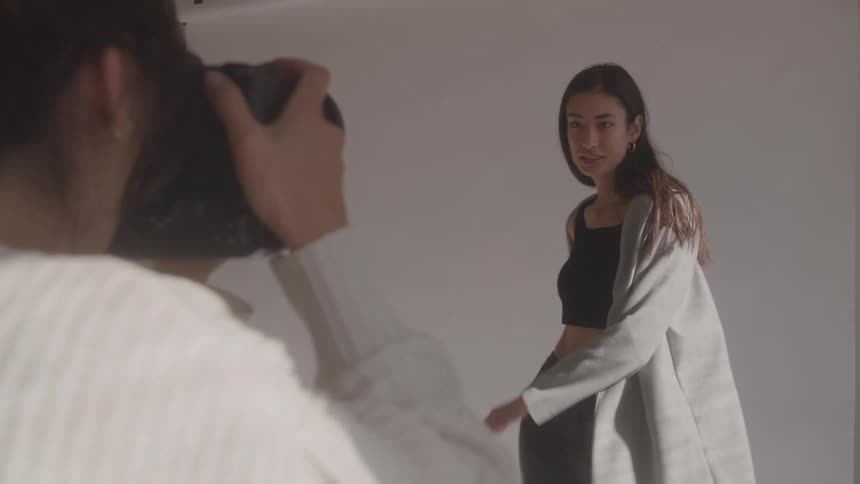 A photographer woman with dark hair is seen from the back taking photos of a young woman model with long dark hair wearing a black crop top and grey jacket as the model poses and smiles against a plain white background