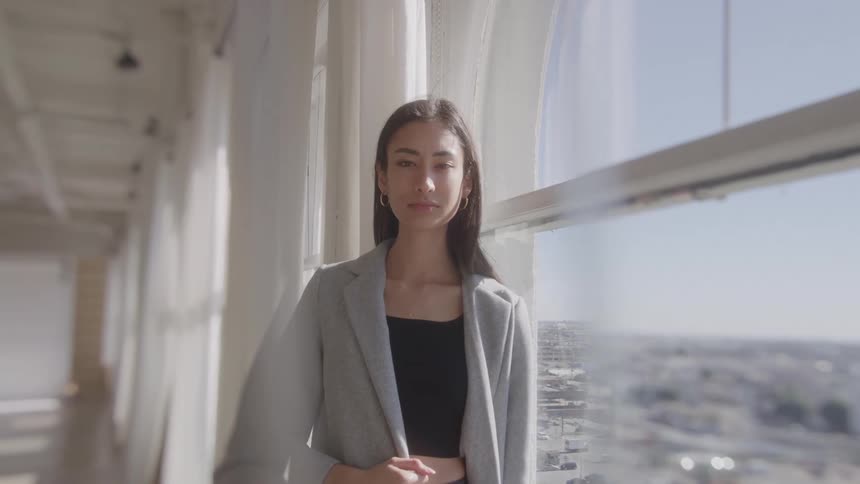 A confident woman in a grey blazer stands near a large window overlooking a city looking directly at the camera