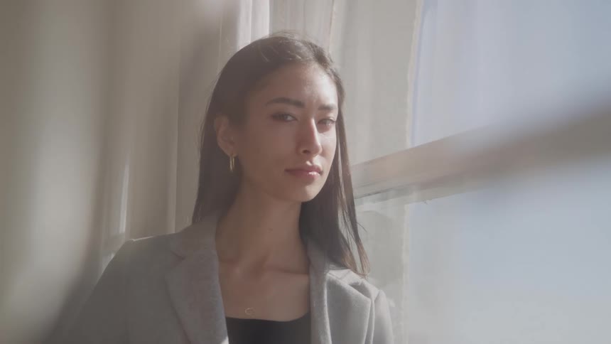 A serious woman with long dark hair standing near a bright window and looking directly at the camera