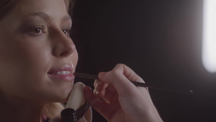 Female makeup artist applying makeup to a female model face with a brush indoors during day time