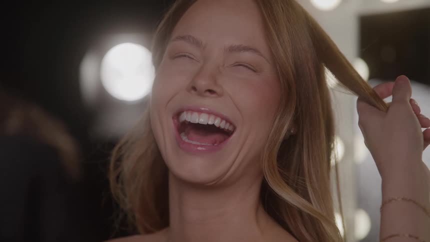 A young woman is in a salon getting her hair done bursting into a huge laugh as the hairdresser styles her hair