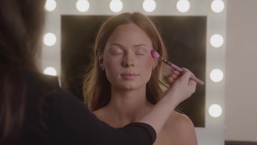 A young woman model with long light brown hair and bare shoulders sits with closed eyes as a makeup artists hand applies blush with a pink brush in front of a vanity mirror with bright lights