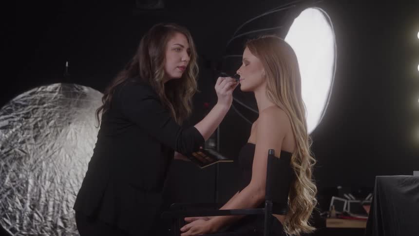 A makeup artist wearing a dark jacket stands over a model seated in a chair applying makeup to the models face with a thin brush