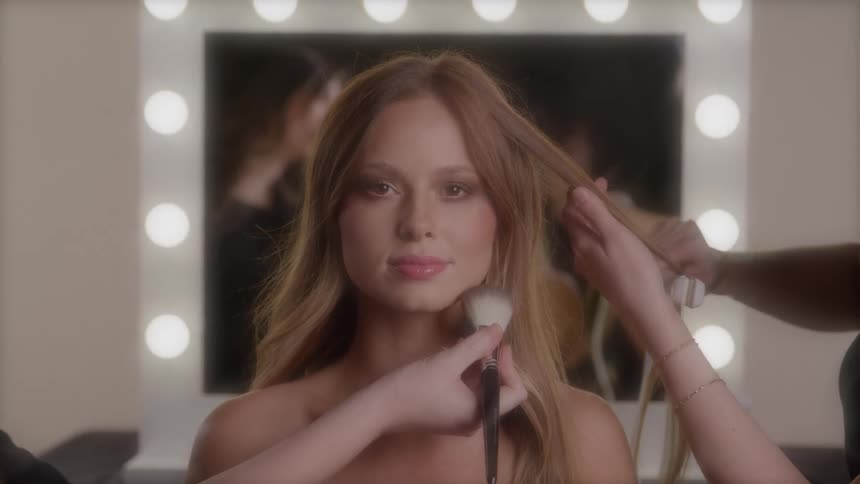 Woman getting makeup and hair done simultaneously in a lit dressing room