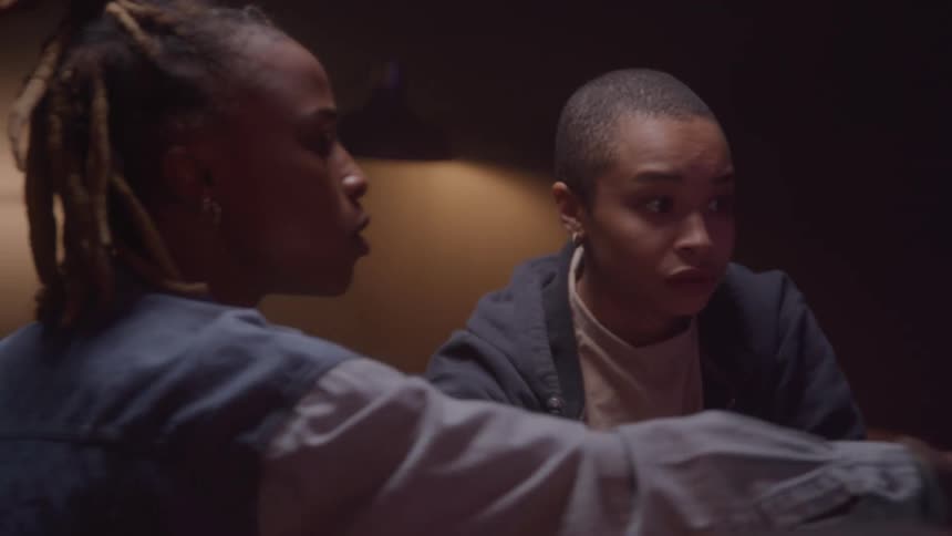 Two young black women sit indoors engaged in a discussion
