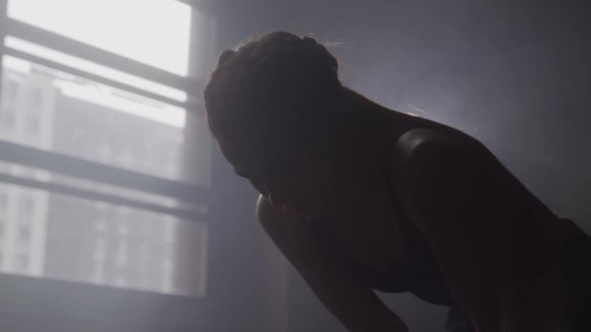Silhouetted woman leans forward in hazy studio light catching her breath after an intense workout