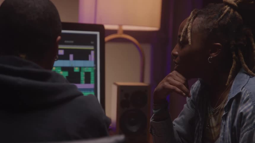 Close up of a young black man and woman discussing while working with a desktop computer at a table in a music studio
