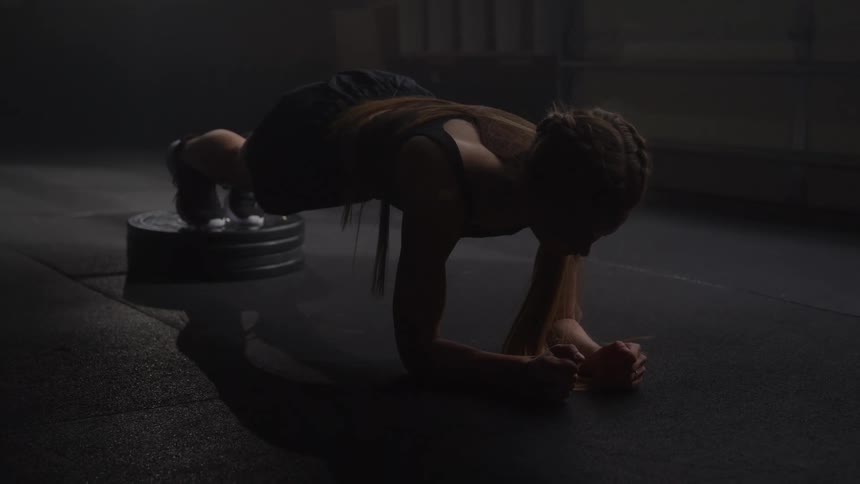 A woman holds an elevated foot plank in a dim gym expression focused and determined