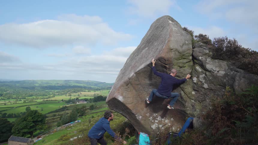 Man climbs a large boulder with a spotter below outdoors